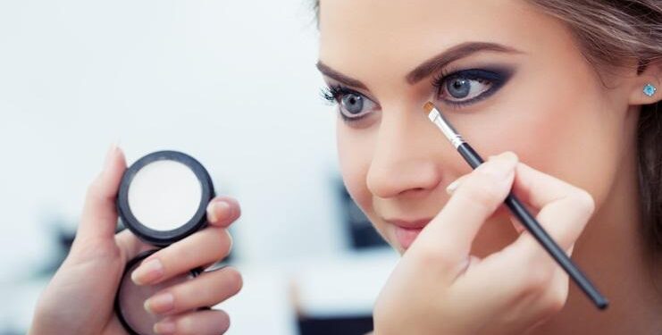 Make-up artist applying white eyeshadow in the corner of model's eye and holding a shell with eyeshadow on background, close up